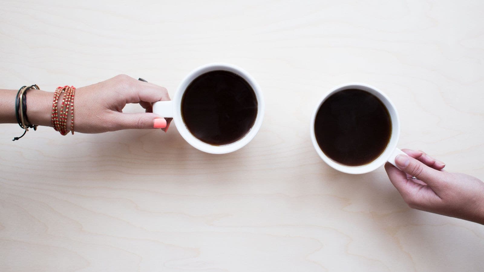 Zwei Hände halten Kaffeetassen über einem hellen Holztisch, der Beginn eines ruhigen Gesprächs