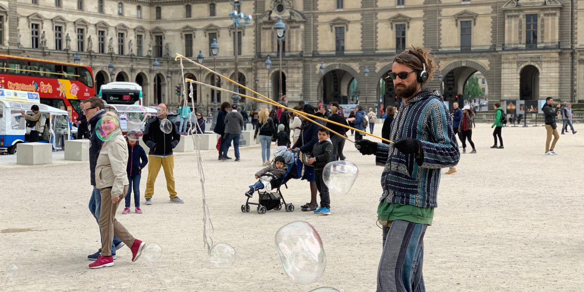 Un artista di strada con le cuffie crea enormi bolle di sapone in una piazza affollata davanti al Louvre, a Parigi