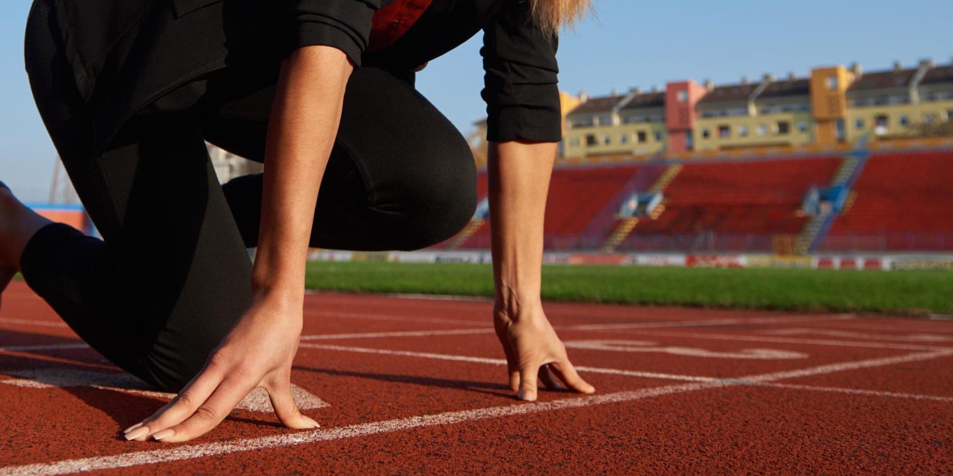 Una velocista accovacciata sulla linea di partenza di una pista rossa, con la punta delle dita sulla linea bianca e uno stadio vuoto alle spalle