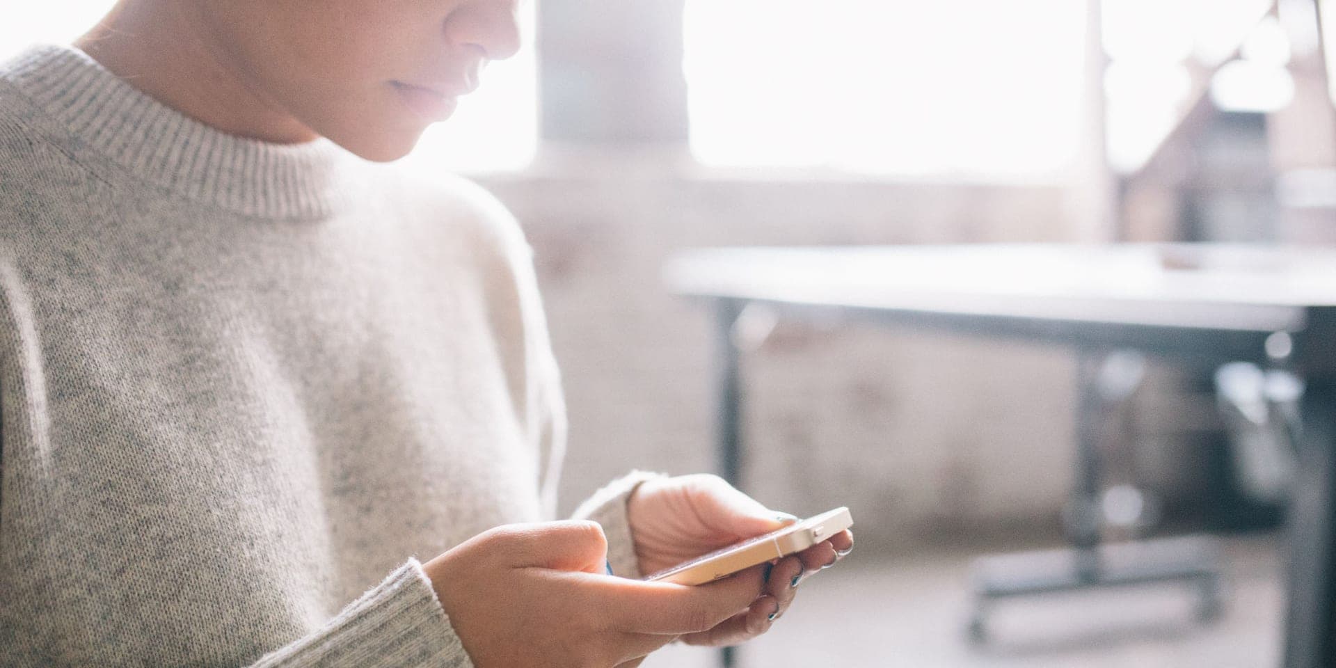 A person in a gray knit sweater looking down at a phone held in both hands, in a bright room with a table behind them